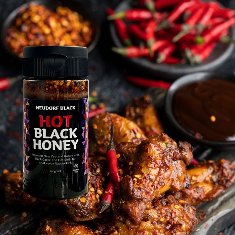 Neudorf Black Hot Black Honey squeezy bottle with roasted chicken wings and red peppers on a dark surface