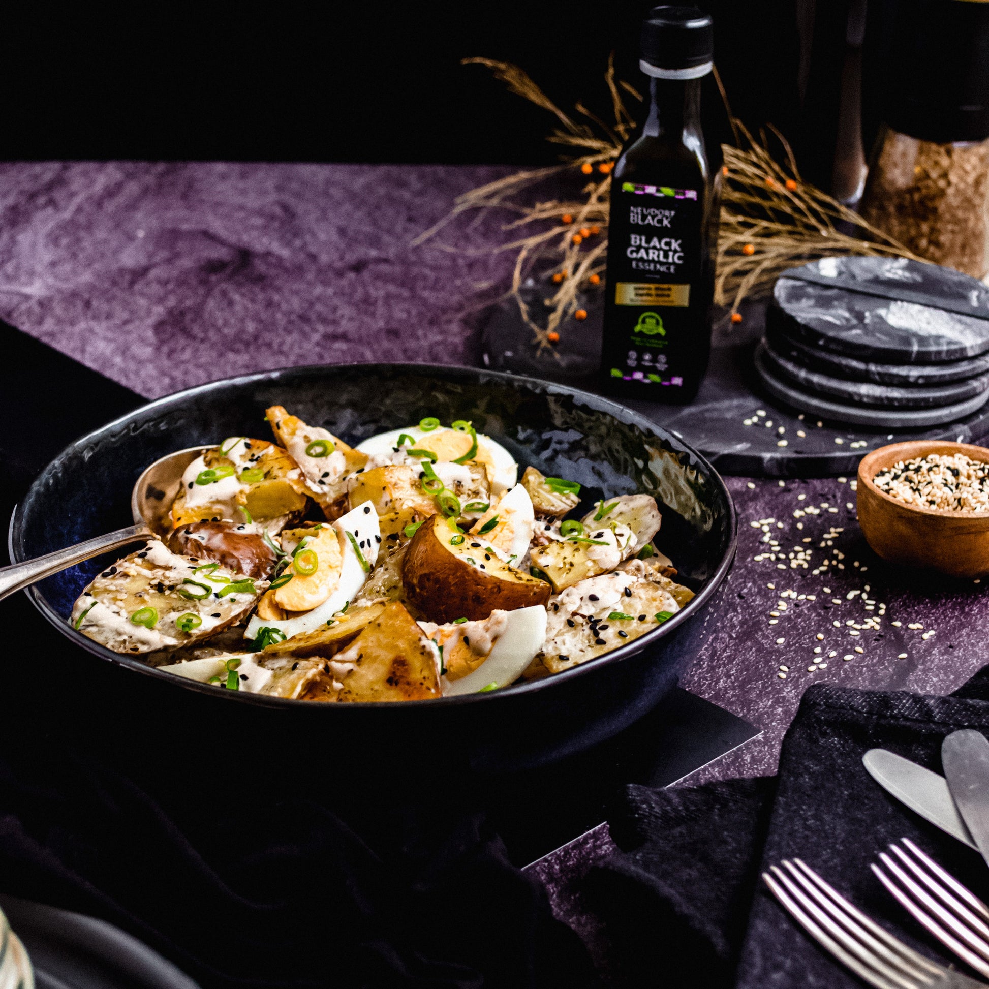 Black Garlic Essence 100ml bottle next to a salad of eggs and roast potatoes that it has been drizzled over