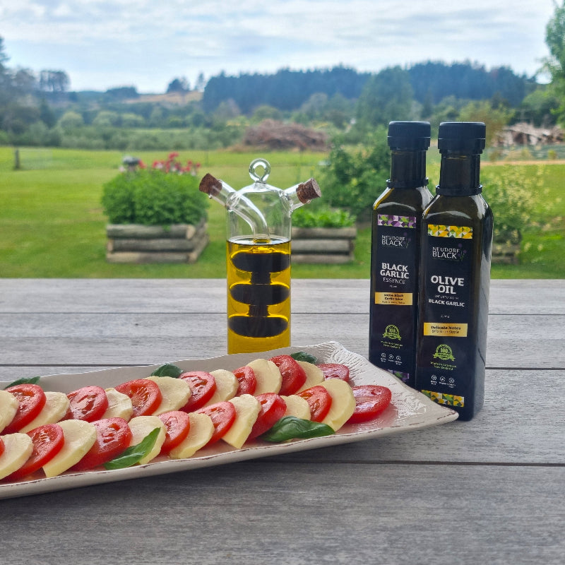 A bottle of Neudorf Black Garlic olive oil beside tomatoes and cheese on a wooden table.