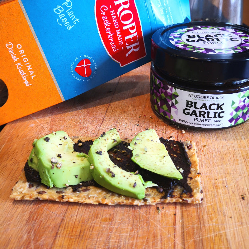 Cracker with avocado slices topped with black garlic puree, next to a jar of black garlic puree and a box of crackers.