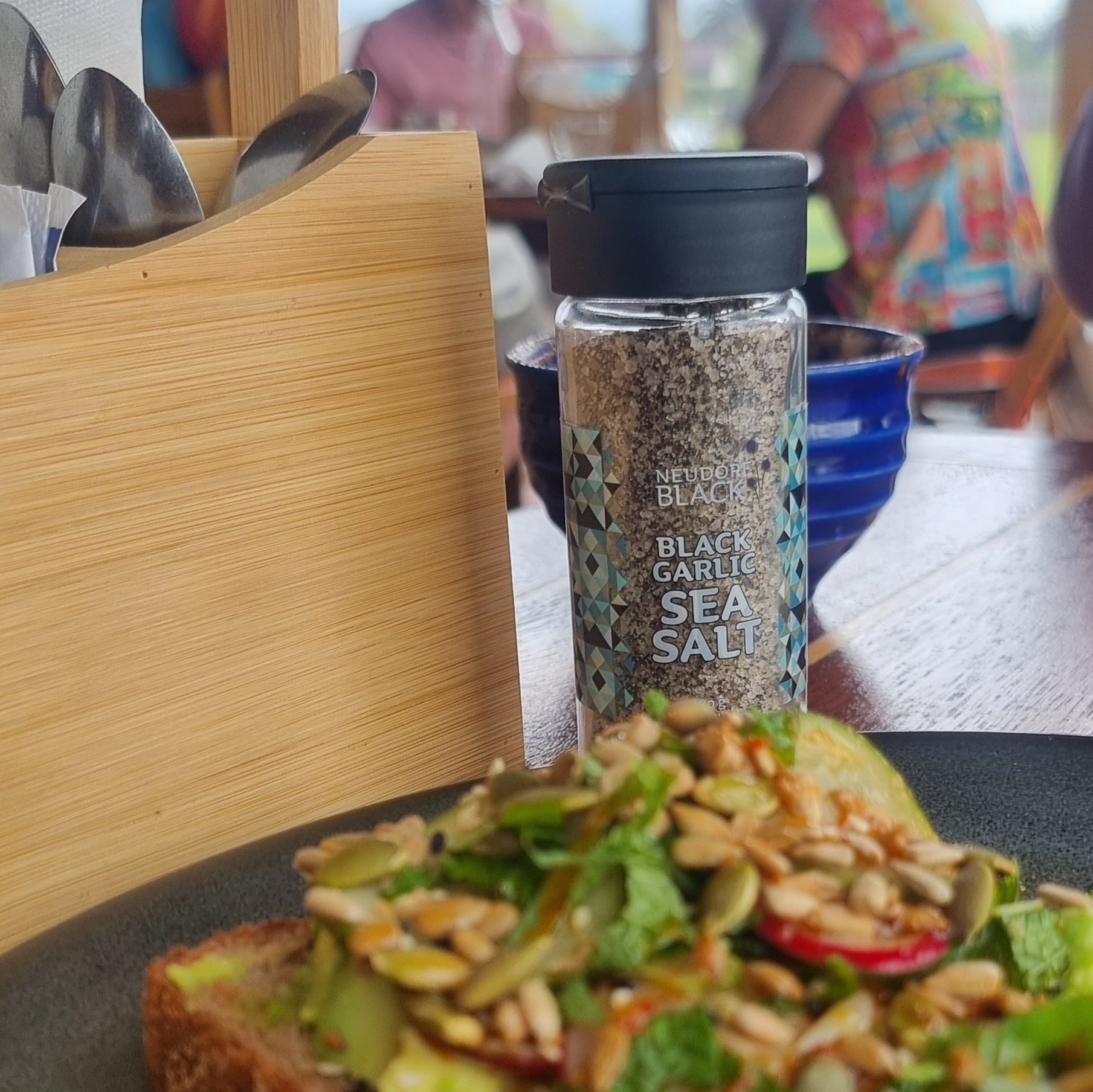 Neudorf Black Garlic Sea Salt in reusable 80g glass shaker beside avocado toast - Marlborough Sea Salt blended with black garlic powder