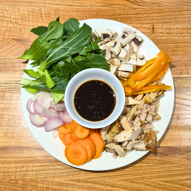 Fresh vegetables and protein arranged on a plate, ready to stir-fry with a bowl of black garlic stir fry sauce sitting on top, ready to use