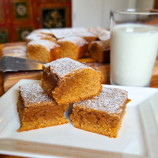 Gingerbread brownies sprinkled with icing sugar on a plate with a cup of milk beside.