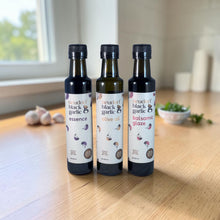 Three bottles of 'Neudorf black garlic' condiments on a wooden table: 'essence', 'olive oil', and 'balsamic glaze'.