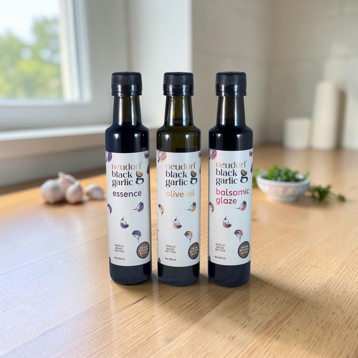Three bottles of 'Neudorf black garlic' condiments on a wooden table: 'essence', 'olive oil', and 'balsamic glaze'.