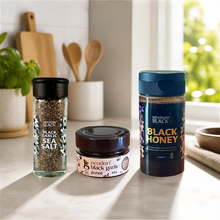 Three gourmet condiments from Neudorf Black are arranged on a kitchen counter in natural sunlight: a glass jar of black garlic sea salt, a small jar of black garlic puree, and a container of black honey.