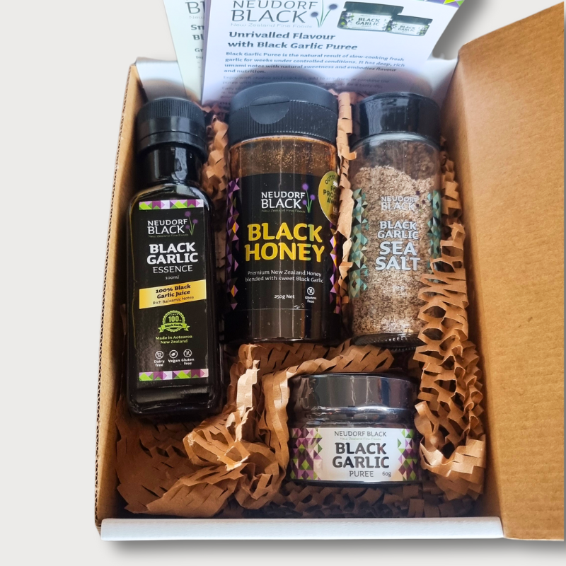 An open cardboard gift box displaying Neudorf Black Garlic bottles and containers of Black Garlic Sea Salt Shaker, Black Honey, Black Garlic Puree, and Black Garlic Essence, arranged on brown paper bedding.