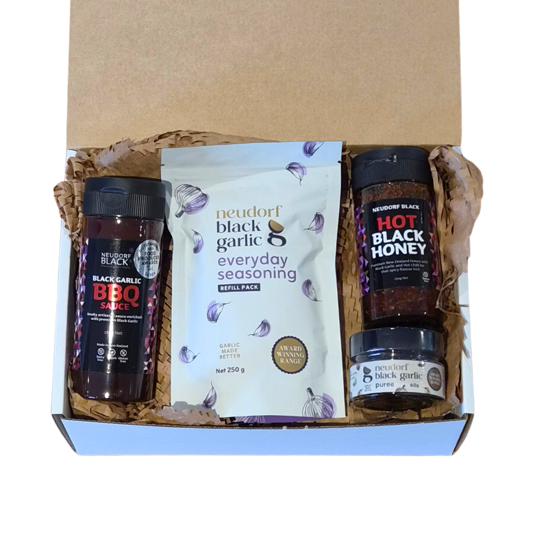 A gift box set containing Neudorf Black Garlic BBQ Sauce, Hot Black Honey, and Everyday Seasoning, arranged in brown paper shred inside a white and brown cardboard box.