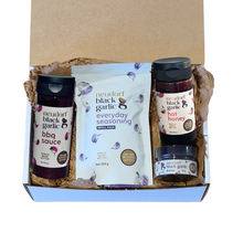 A Neudorf Black Garlic gift set in a cardboard box with brown paper shred. The set includes a bottle of Neudorf Black Garlic BBQ Sauce, a bag of Neudorf Black Garlic Everyday Seasoning, a jar of Neudorf Black Garlic Hot Honey, and a small jar of Neudorf Black Garlic puree