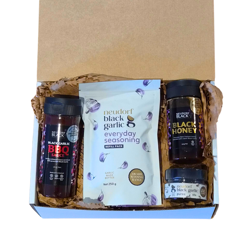 A gift box set containing Neudorf Black Garlic BBQ Sauce, Black Honey, and Everyday Seasoning, arranged in brown paper shred inside a white and brown cardboard box.