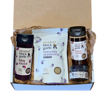 A gift box set of Neudorf Black Garlic products arranged in brown paper shred. The set includes a bottle of black garlic BBQ sauce, a bag of everyday seasoning, a jar of black garlic honey, and a small jar of black garlic puree.