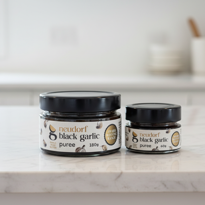 Two jars of Neudorf Black Garlic Puree on a white marble countertop in a bright, modern kitchen. The larger jar is 180g and the smaller jar is 60g, both with black lids and white labels featuring black and gold text.