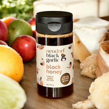 A jar of Neudorf Black Garlic Black Honey centered on a wooden surface, surrounded by food props including apples, bread, cheese, and citrus. The product label is clearly visible, and the jar has a black lid.