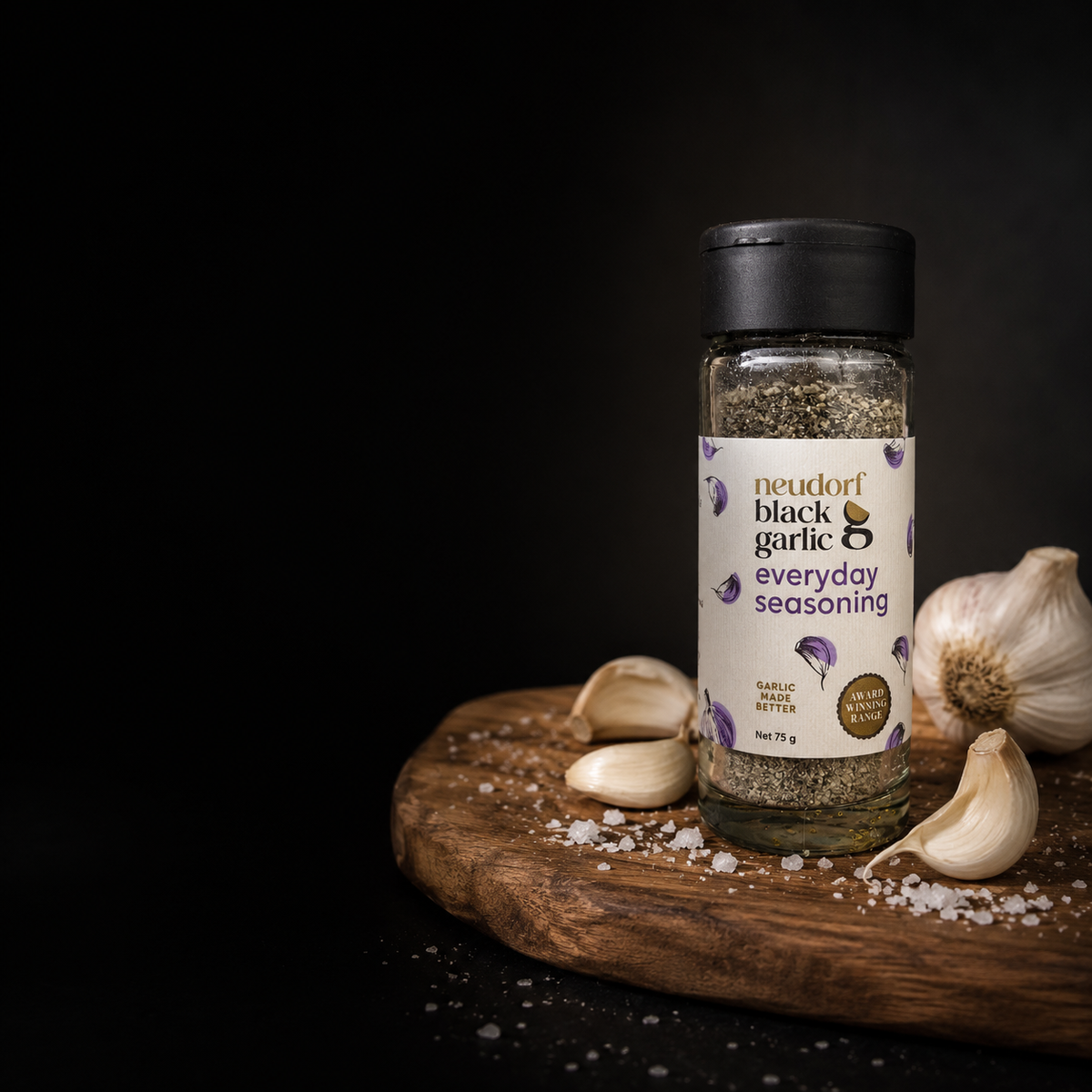 A bottle of Neudorf Black Garlic Everyday Seasoning stands on a rustic wooden board with garlic cloves and coarse salt scattered around it against a dark background.