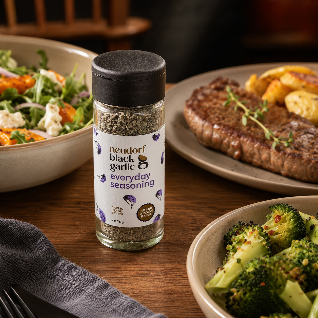 A jar of Neudorf Black Garlic Everyday Seasoning on a wooden table in a dining setting, surrounded by a steak, broccoli, and salad.