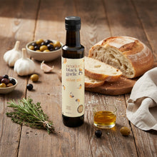 A bottle of Neudorf Black Garlic Olive Oil stands on a rustic wooden table, surrounded by bread, garlic, olives, fresh thyme, and a small glass of the golden oil.