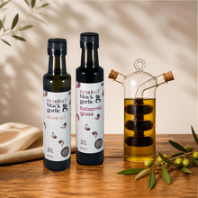 Two bottles of Neudorf Black Garlic products on a wooden table: a bottle of olive oil and a bottle of balsamic glaze, next to a glass carafe filled with the oil and balsamic, with olive branches in the background and foreground.