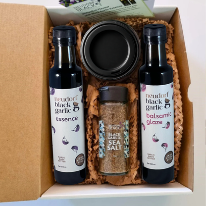 A gift box set containing two bottles of Neudorf Black Garlic products, an essence bottle and a balsamic glaze bottle, a jar of black garlic sea salt, and a jar of puree, arranged in brown paper shred inside a cardboard box.