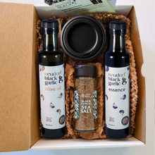 A gift box set containing two bottles of Neudorf Black Garlic products, an olive oil bottle and an essence bottle, a jar of black garlic sea salt, and a jar of puree, arranged in brown paper shred inside a cardboard box.