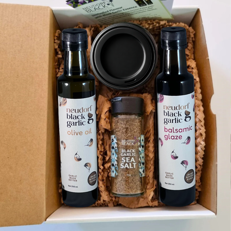 A gift box set containing two bottles of Neudorf Black Garlic products, an olive oil bottle and a balsamic glaze bottle, a jar of black garlic sea salt, and a jar of puree, arranged in brown paper shred inside a cardboard box.